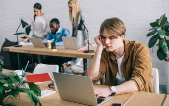 A young man wearing a brown jacket sits in front of a laptop with his head resting on his hand, appearing bored or tired. In the background, three colleagues work at a table with laptops and papers, surrounded by potted plants in a white modern office.