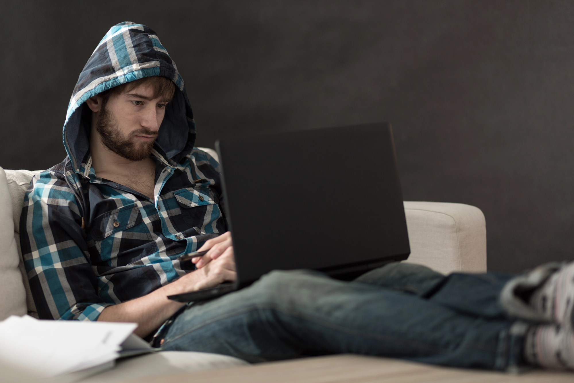 A man in a hooded plaid shirt and jeans is sitting on a sofa with his legs extended on a wooden table. He is focused intently on a large laptop resting on his lap. Papers are scattered on the table beside him, and the background is a dark, plain wall.