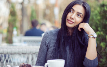 A woman with long black hair sits at an outdoor café, resting her head on her hand and smiling thoughtfully. She wears a grey sweater and a watch. In front of her is a white cup and a slice of chocolate cake. The background is blurred with greenery and other patrons.