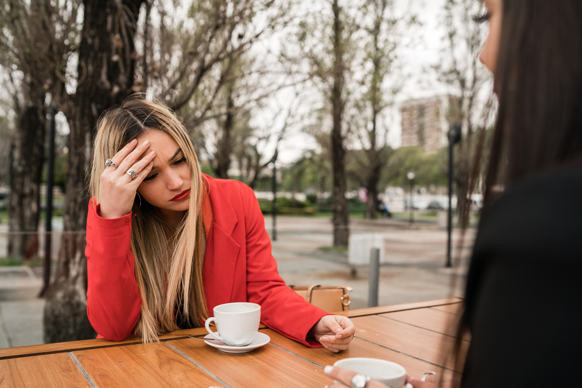 A woman wearing a red jacket sits at an outdoor table with a cup of coffee, holding her head as if she has a headache. Another person, slightly out of focus, sits across from her. The background shows trees and a park-like setting.