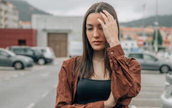 A young woman in a brown jacket stands in a parking lot with her head slightly tilted and her hand touching her hair. She appears to be in deep thought. The background includes parked cars and buildings under a cloudy sky.