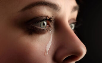 Close-up of a woman's face with a single tear rolling down her cheek. The image focuses on her expressive eyes with smeared eye makeup, reflecting an emotion of sadness or distress. The background is dark, highlighting her facial features.