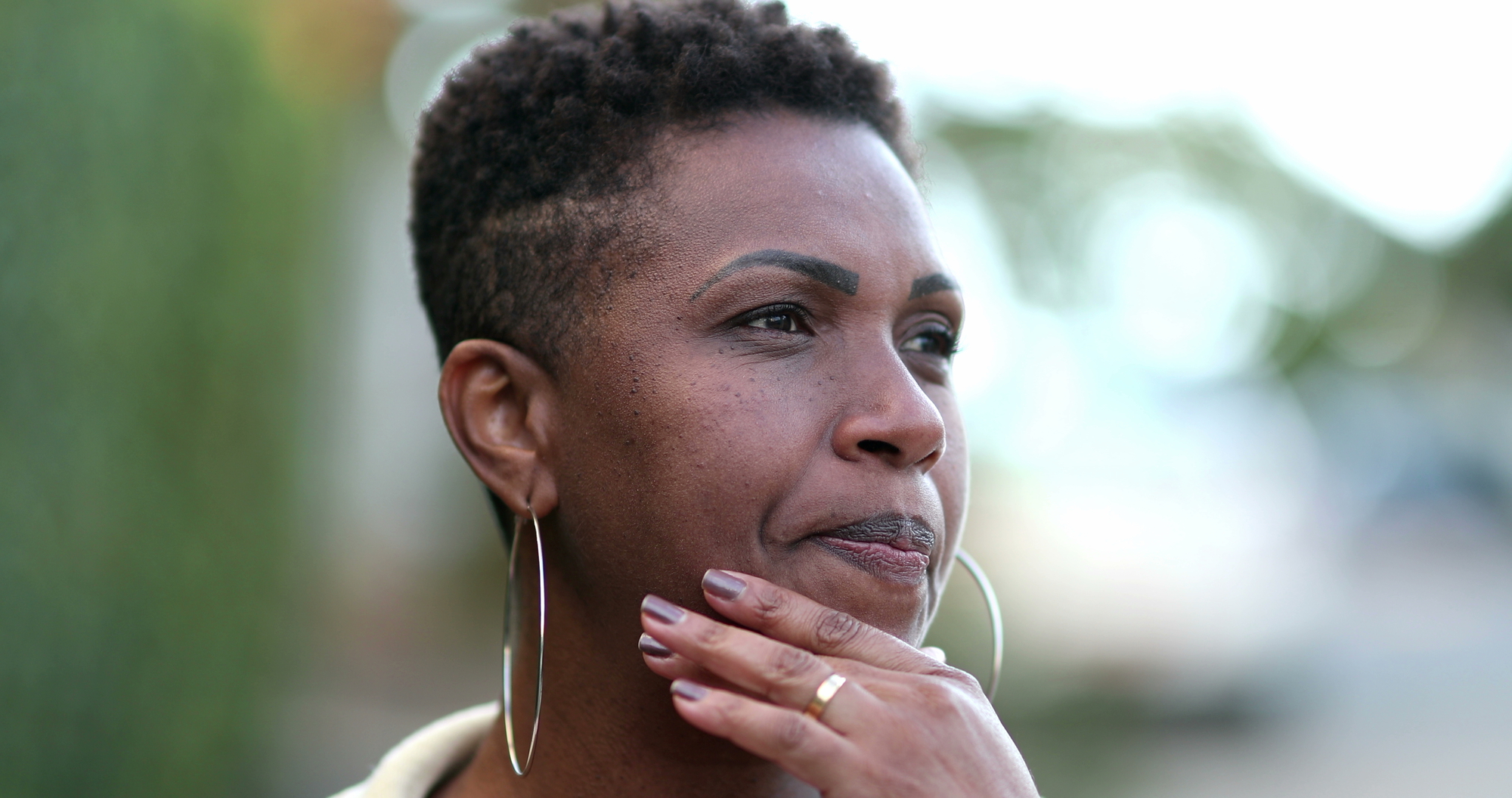 A woman with short curly hair is looking pensively to the side, with her hand touching her chin. She is wearing hoop earrings and has a thoughtful expression. The background is softly blurred.