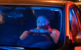 A person with an afro hairstyle is seen through the windshield of a car at night. They appear concerned, gripping the steering wheel tightly. The scene is illuminated by blue and red lights, suggesting the presence of emergency or police vehicles nearby.