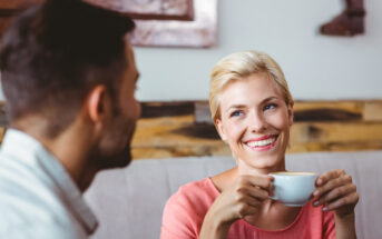 A smiling woman with short blond hair holds a white coffee cup while looking at a man with dark hair who is in profile. They are sitting in a cozy café with wooden decor and enjoying a conversation.