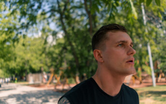 A young man with short hair and a tattoo on his left arm is standing outdoors, looking to the side with a thoughtful expression. He is wearing a black sleeveless shirt. The background is a sunlit park with trees, grass, and a dirt path.