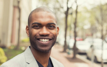 A smiling man with a shaved head and well-groomed beard is wearing a light gray blazer. He is standing outdoors on a sidewalk, with out-of-focus buildings and trees in the background. The overall scene appears bright and lively.