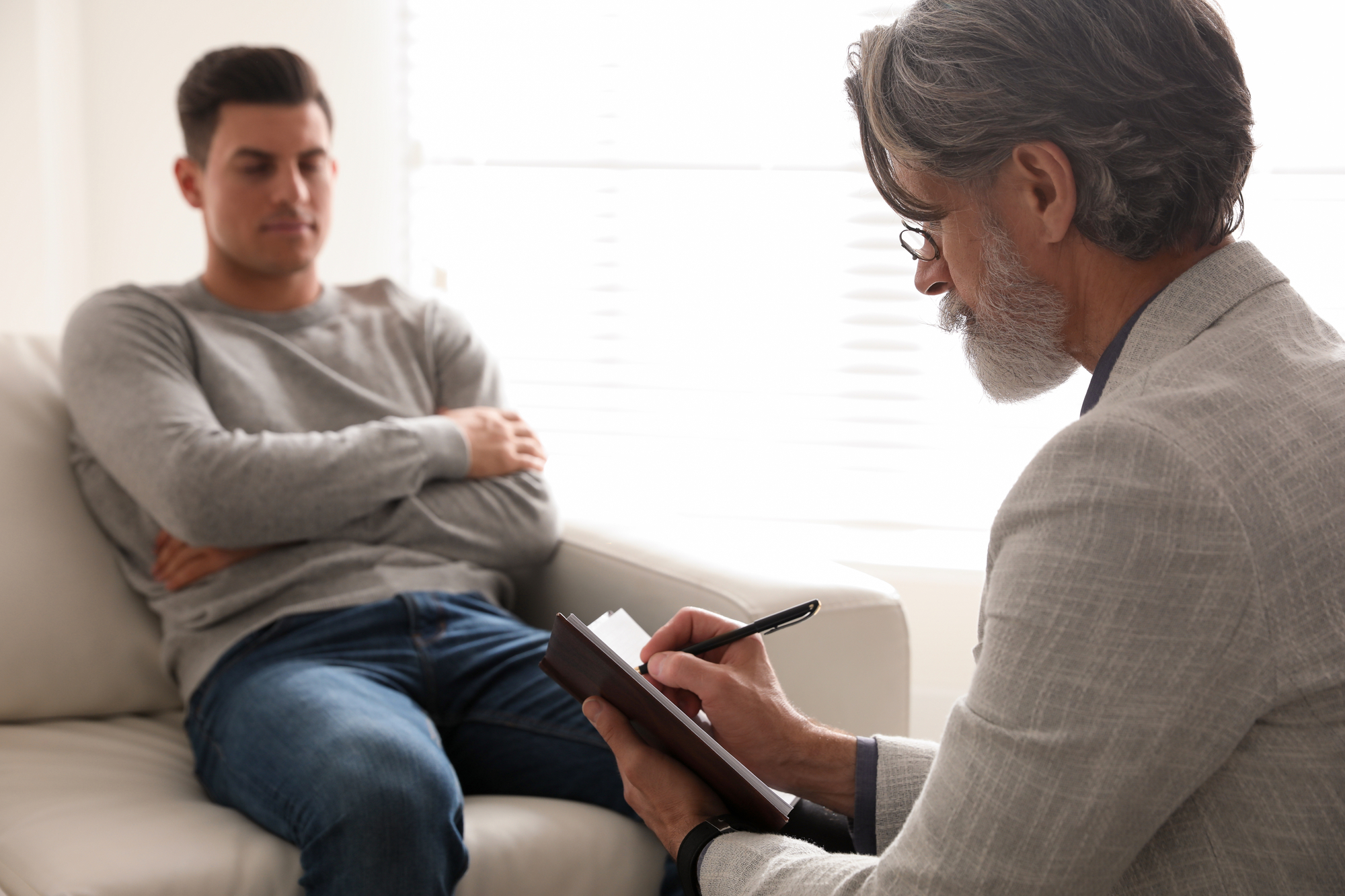 A man with folded arms sits on a couch, looking reflective, while another man with graying hair and glasses takes notes on a clipboard. The setting appears to be a bright, professional office, suggesting a therapy or counseling session.