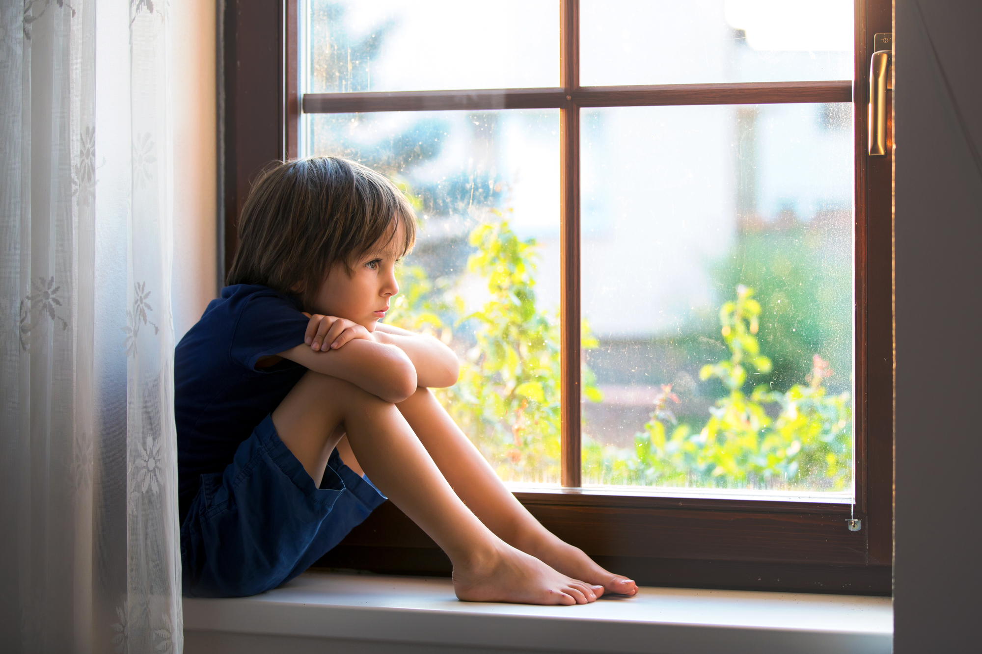 A young boy with brown hair sits on a window sill, hugging his knees and looking outside. He is wearing a blue shirt and shorts. Sunlight filters through the window, illuminating the room and casting a soft glow. There are sheer curtains to the left.