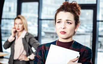 A woman with curly hair and a concerned expression holds a clipboard while standing in the foreground. In the background, another woman with blonde hair and a serious expression talks on the phone. They are in a modern office space with large windows.