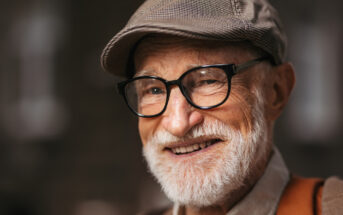 An older man with a white beard and glasses is smiling warmly. He is wearing a flat cap and a brown jacket. The background is blurred, focusing attention on his joyful expression.