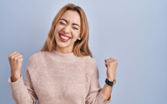 A young woman with long blonde hair is standing against a light blue background. She is smiling widely with her eyes closed and her fists clenched, expressing joy and excitement. She is wearing a beige sweater and a watch on her left wrist.