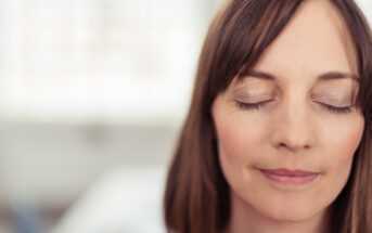 A woman with shoulder-length brown hair and bangs is shown in a close-up with her eyes closed and a serene expression on her face. The background is out of focus, suggesting she might be indoors.