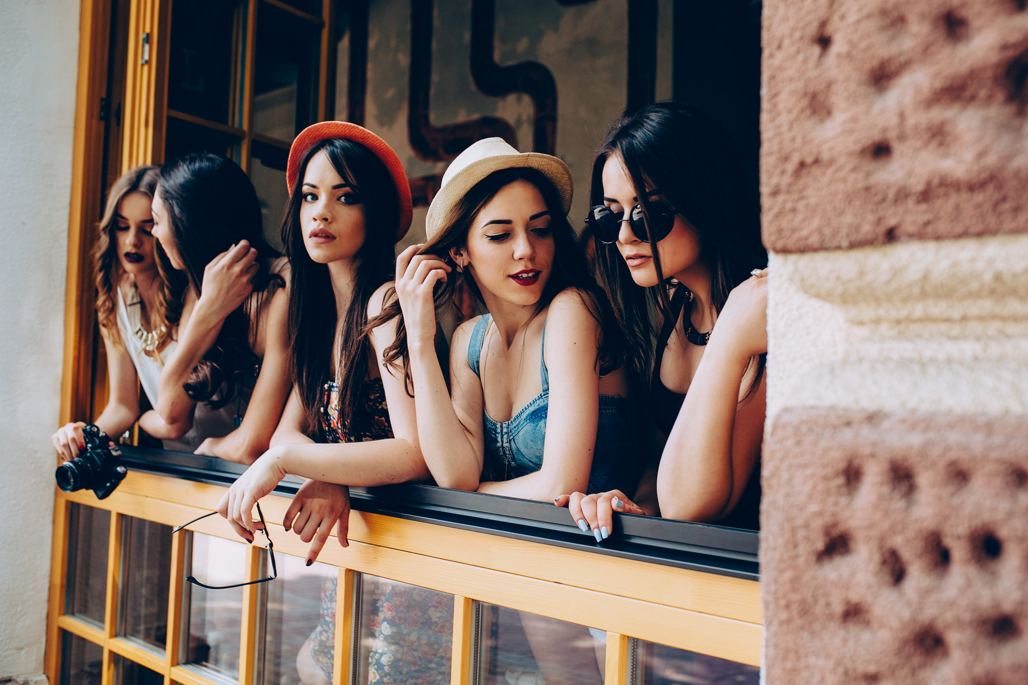 Five women are leaning against a window frame, looking outwards. They are wearing fashionable summer outfits, including hats and sunglasses. The scene appears casual and relaxed, with some women resting their hands on the window ledge.