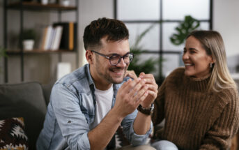 A young man and woman are sitting on a couch, smiling and laughing. The man, wearing glasses and a denim shirt over a white t-shirt, holds something in his hands. The woman, dressed in a brown sweater, looks at him joyfully. A modern home interior is in the background.