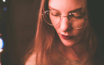 Close-up of a woman with long hair, wearing round glasses. She is looking down, with a serious or thoughtful expression. The lighting is soft and warm, casting a gentle glow on her face. The background is dark and blurred.