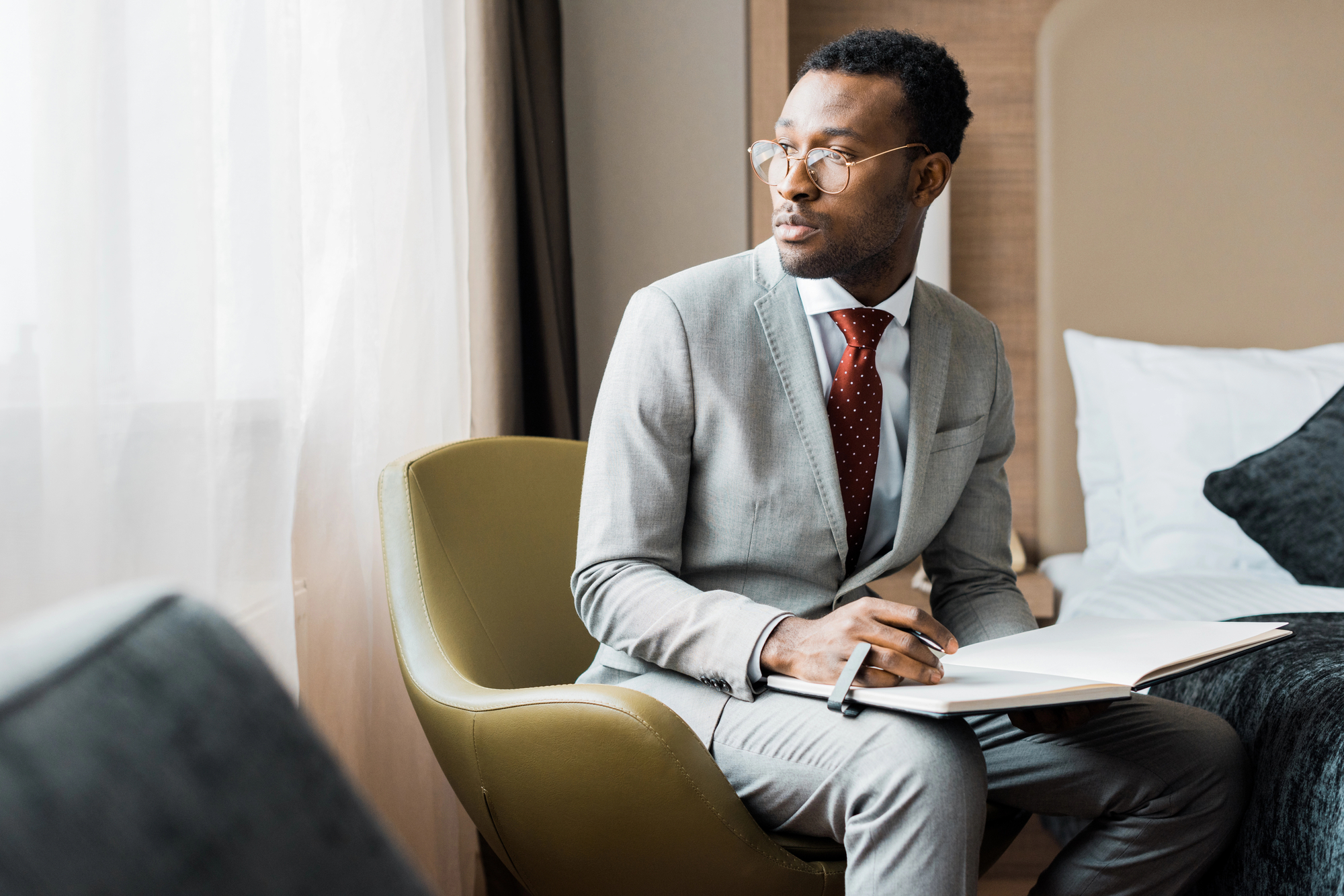 A man in a gray suit with a red tie and glasses sits in a chair by a window, holding an open book and a pen. He appears to be deep in thought and is looking out the window with a contemplative expression. Behind him is a neatly made bed.