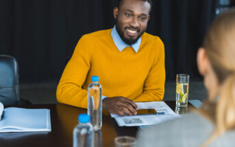 A person in a yellow sweater is sitting at a table with documents, a smartphone, and two water bottles. They are smiling and looking at another individual with blonde hair seated across from them. The setting appears to be a professional or interview environment.