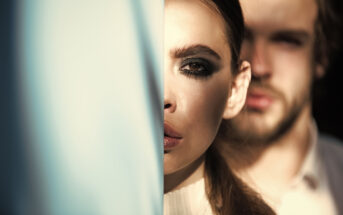 Close-up of a woman partially obscured by a blue object, leaving only one heavily made-up eye visible. Behind her, a man with a beard is blurred and partially visible. The lighting casts a dramatic shadow across both faces.