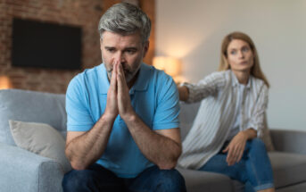 A man with gray hair and a beard sits on a couch with his hands clasped together, looking distressed. A woman with long hair sits next to him, reaching out a hand towards him with a concerned expression. The background shows a living room setting.