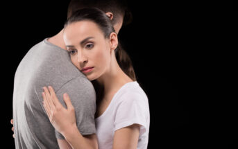 A woman in a white shirt embraces a man in a gray t-shirt from behind, resting her head on his shoulder. They are against a black background, and the woman has a contemplative expression. The man's face is not visible.