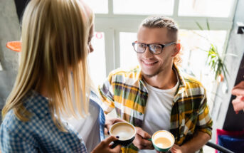 Two people in casual attire enjoying coffee together indoors. One person with glasses and a plaid shirt is smiling at the other person, who has long blonde hair and is wearing a blue checkered shirt. They are standing near a window with natural light streaming in.