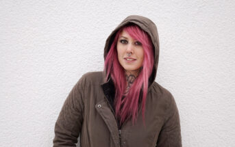 A person with pink hair wearing a brown hooded jacket smiles at the camera. They have several facial piercings and visible tattoos on their neck. The background is a plain, textured white wall.