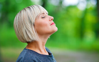 A woman with short blonde hair and closed eyes stands in a green outdoor setting, appearing to enjoy the fresh air. She is wearing a dark-colored top, and the background is blurred with lush foliage.