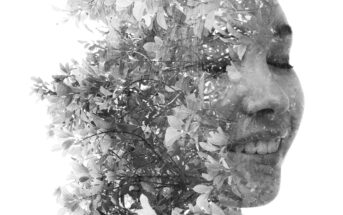 A double exposure black-and-white image combines the profile of a smiling person with closed eyes and overlapping foliage. The person's face is subtly blended with intricate leaves and branches, creating a serene and harmonious visual effect.