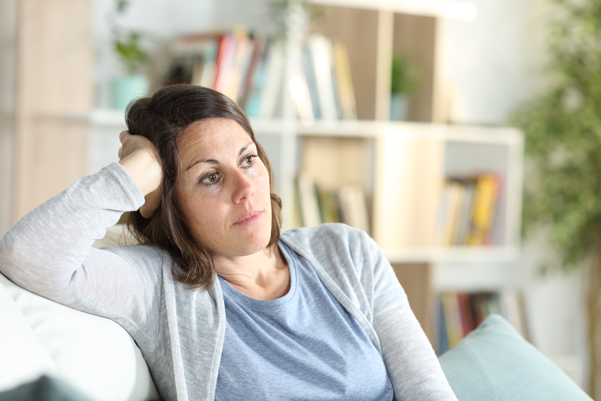 A woman with medium-length brown hair is sitting on a couch, resting her head on her hand. She appears thoughtful or pensive. She is wearing a blue shirt and light gray cardigan. In the background, there is a blurry bookshelf with books and plants.