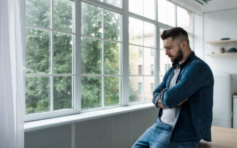 A man with a beard and short hair stands pensively by a large window with greenery visible outside. He is wearing a denim jacket over a white shirt and jeans, with his arms crossed and eyes looking down. The interior is bright with natural light from the window.