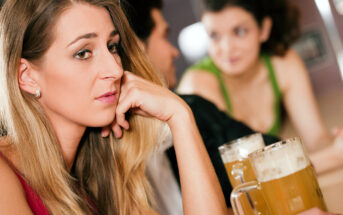 A woman with long blonde hair rests her chin on her hand, looking pensive, at a bar. In the background, a man and a woman with curly hair are engaged in conversation. Two mugs of beer are on the bar counter in front of them.