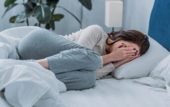 A person is lying on a bed, curled up with their hands covering their face. They are wearing a long-sleeve top and sweatpants. The room has a minimalistic decor with a plant in the background. The atmosphere appears to be calm yet somber.