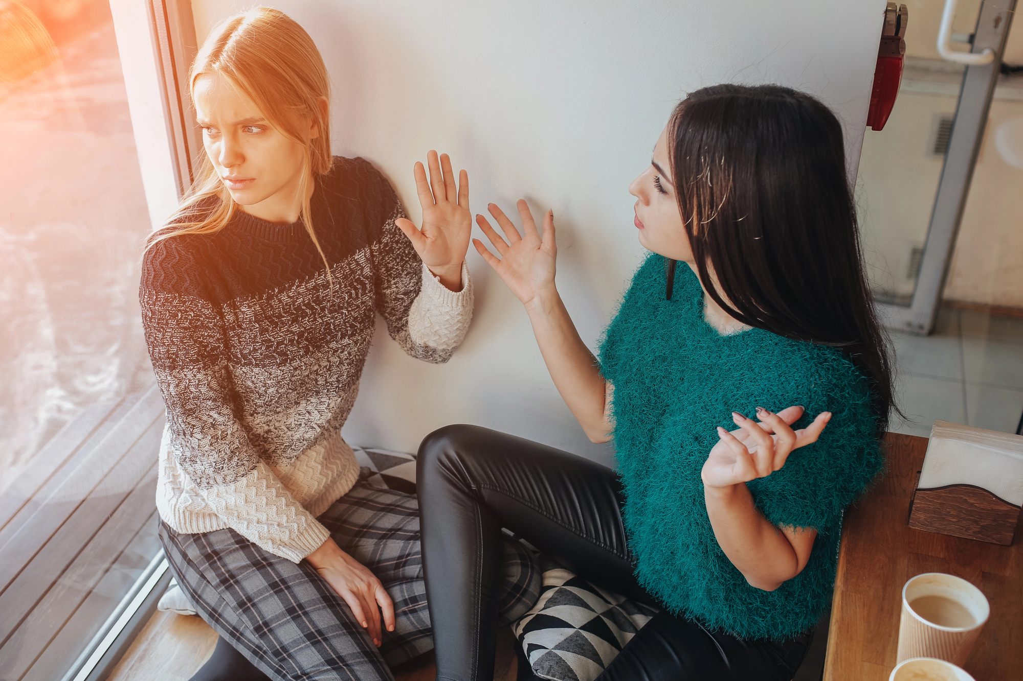 Two women sit on the floor by a window, engaged in an intense conversation. The woman on the left raises her hand as if to stop the other, looking concerned. The woman on the right gestures with both hands, appearing distressed. Two coffee cups sit on the table nearby.