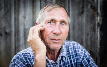 An elderly man with short gray hair and blue eyes, wearing a plaid shirt, looks directly at the camera. He holds his right hand to the side of his face near his eye. The background is a weathered wooden fence.