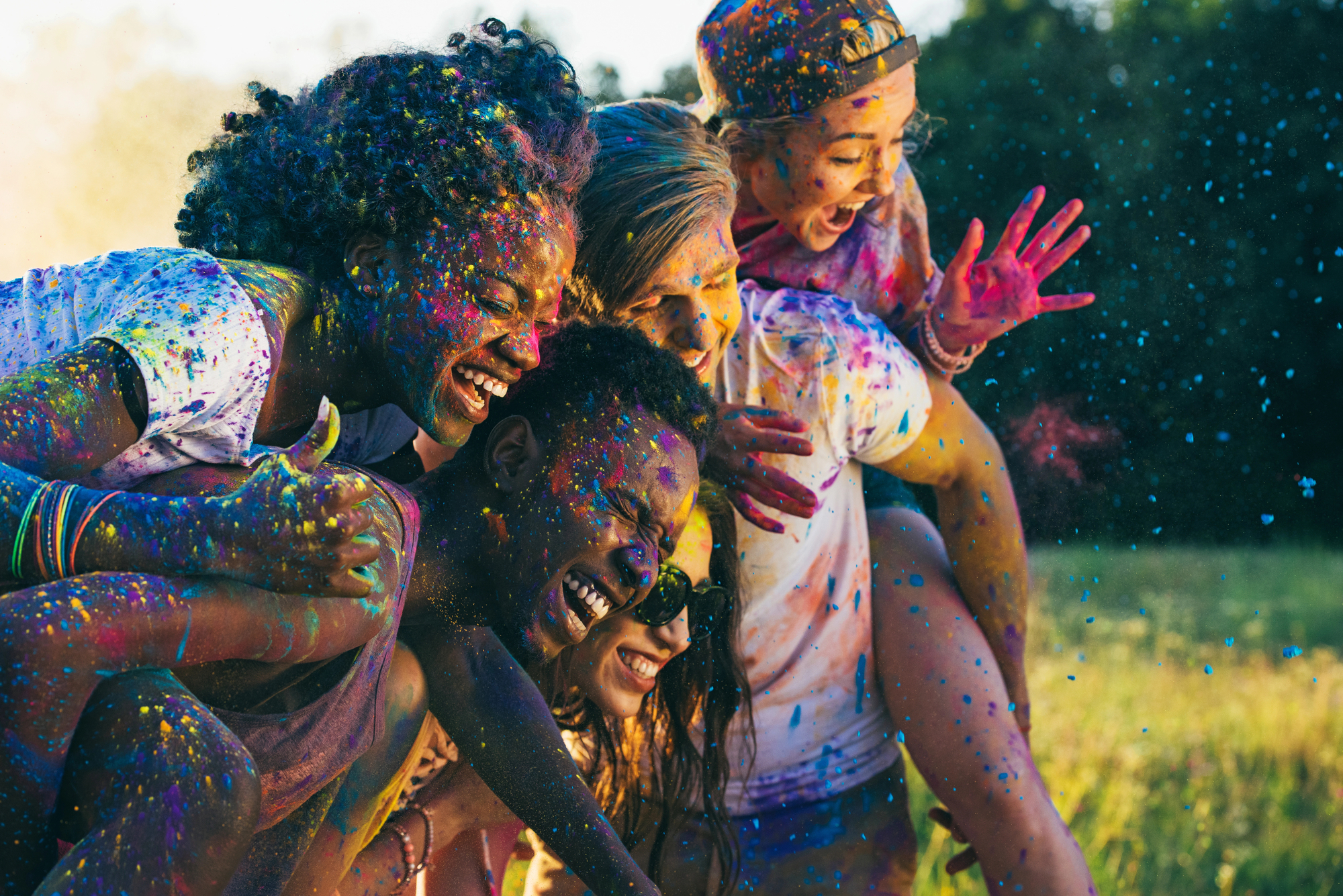 Five people joyfully celebrating with colorful powder on their clothes and faces, outdoors in a sunlit field. They are laughing, with some leaning on each other, radiating happiness and energy. The background features green trees and a bright sky.