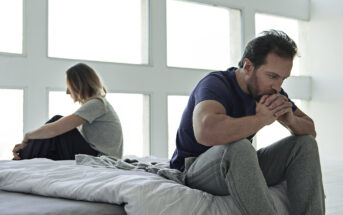 A man and woman sit on a bed facing away from each other. The man in the foreground is leaning forward, resting his elbows on his knees and holding his hands near his mouth, looking pensive. The woman sits in the background hugging her knees, also looking troubled.