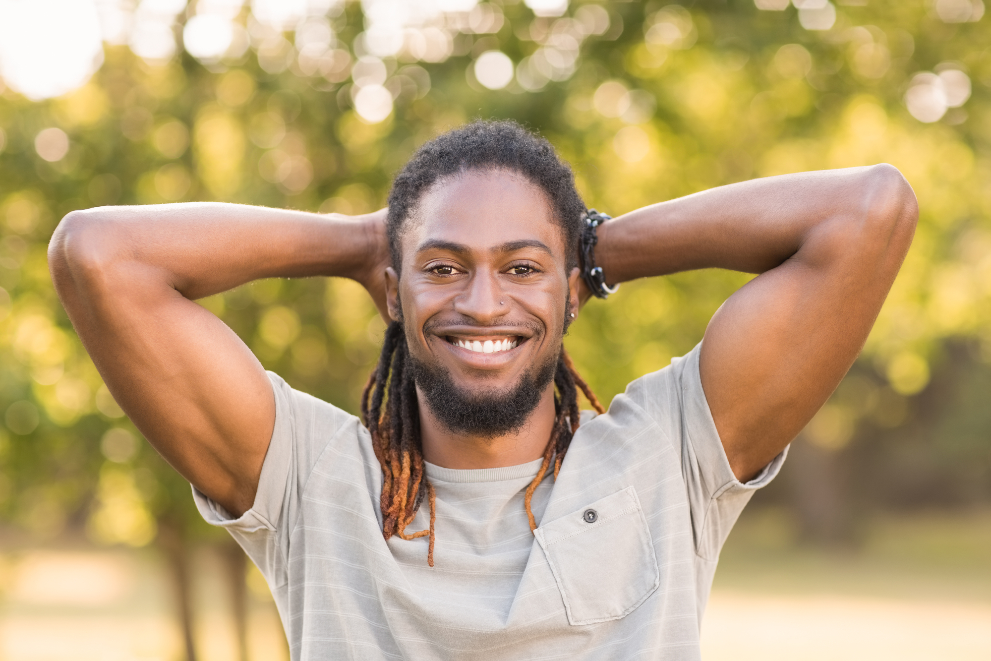 A smiling man with a beard and long, braided hair stands outdoors with his hands resting behind his head. He is wearing a light gray t-shirt, and the background is a sunny, blurred green park scene.