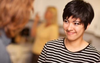 A person with short, dark hair and wearing a black and white striped shirt is smiling warmly at another person, whose back is to the camera. A blurred figure in the background suggests a casual, indoor setting.