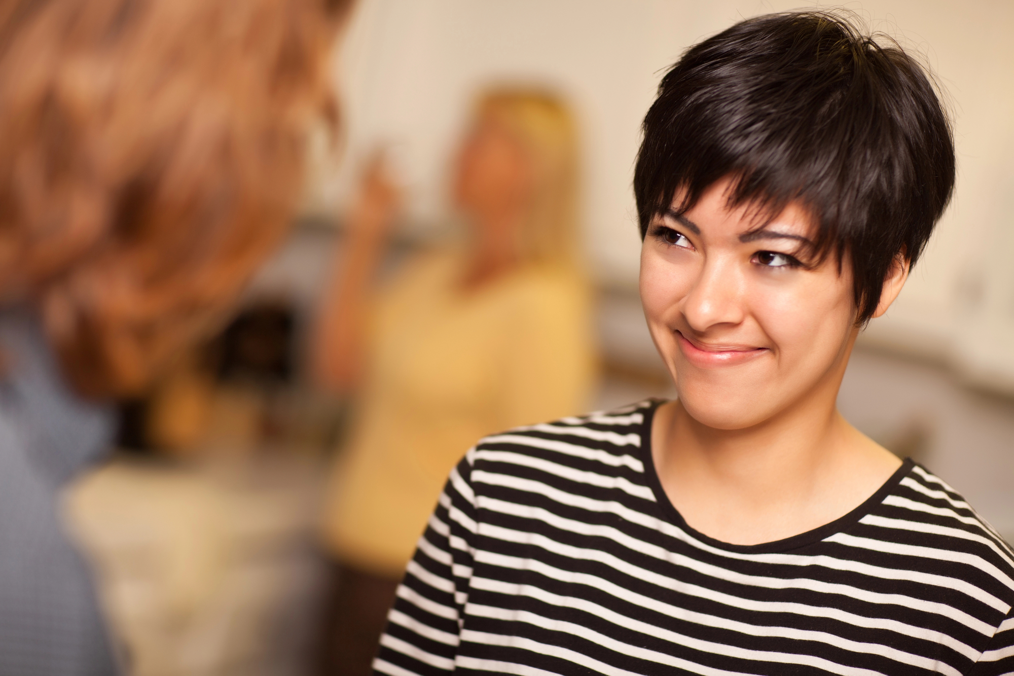 A person with short, dark hair and wearing a black and white striped shirt is smiling warmly at another person, whose back is to the camera. A blurred figure in the background suggests a casual, indoor setting.