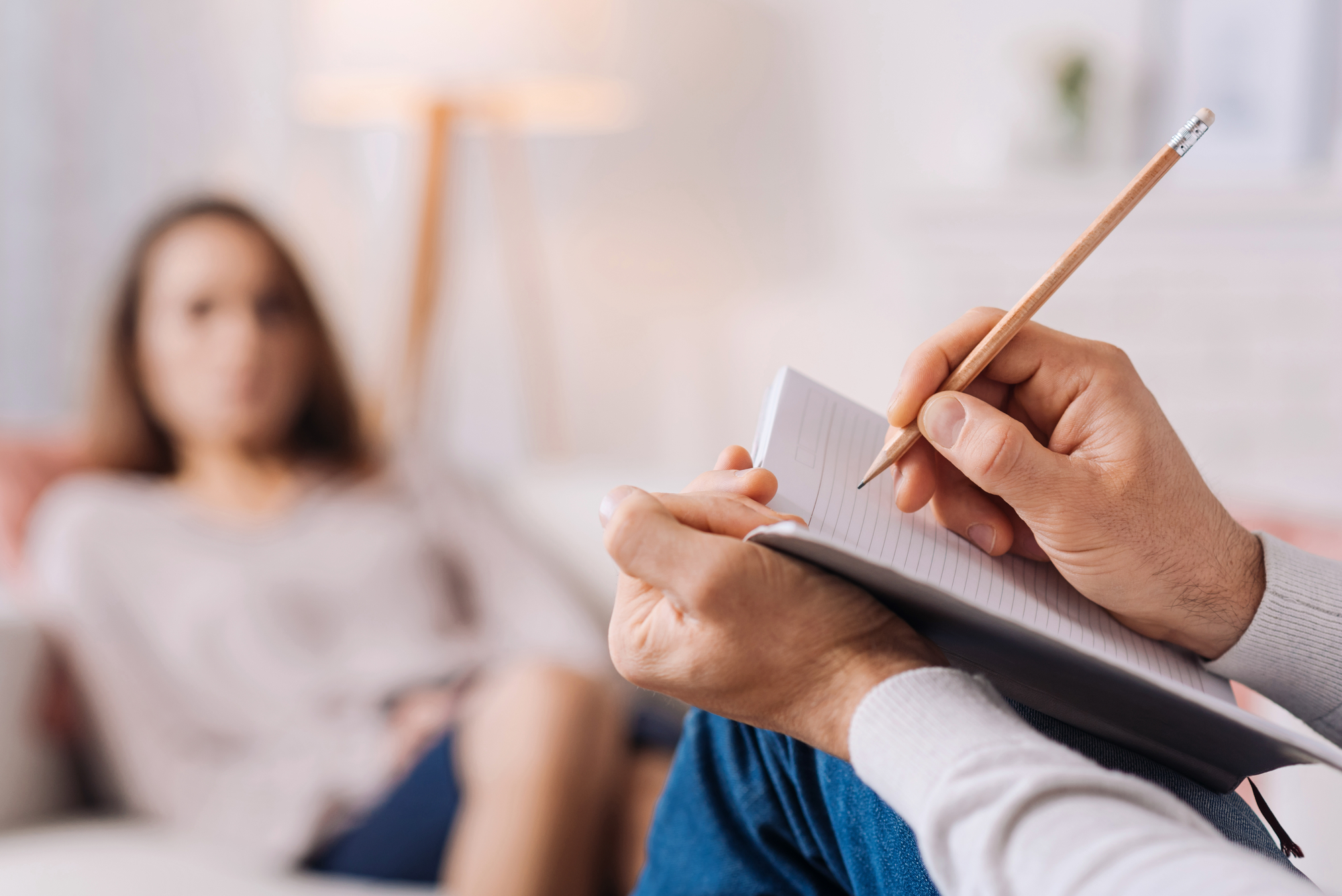 A person sits on a couch, blurred in the background, while another person's hands are in focus in the foreground, holding a notepad and pencil, possibly taking notes during a session, likely in a therapy or counseling setting.