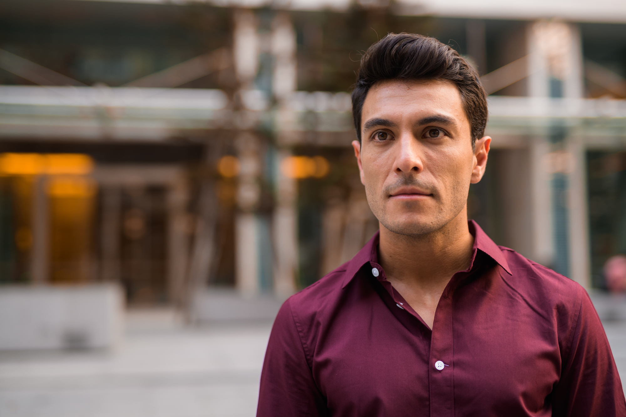 A man with short black hair wearing a maroon button-up shirt stands outside a modern building. He looks into the camera with a neutral expression. The background is blurred, highlighting the architectural elements and greenery behind him.