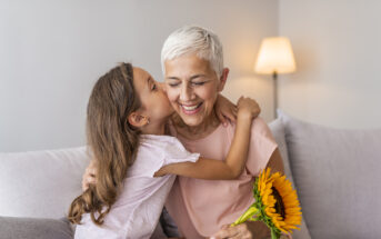 A joyful elderly woman with short white hair is being embraced and kissed on the cheek by a young girl with long brown hair. The woman is smiling and holding a bright sunflower. They are seated on a couch with a lit lamp in the background.