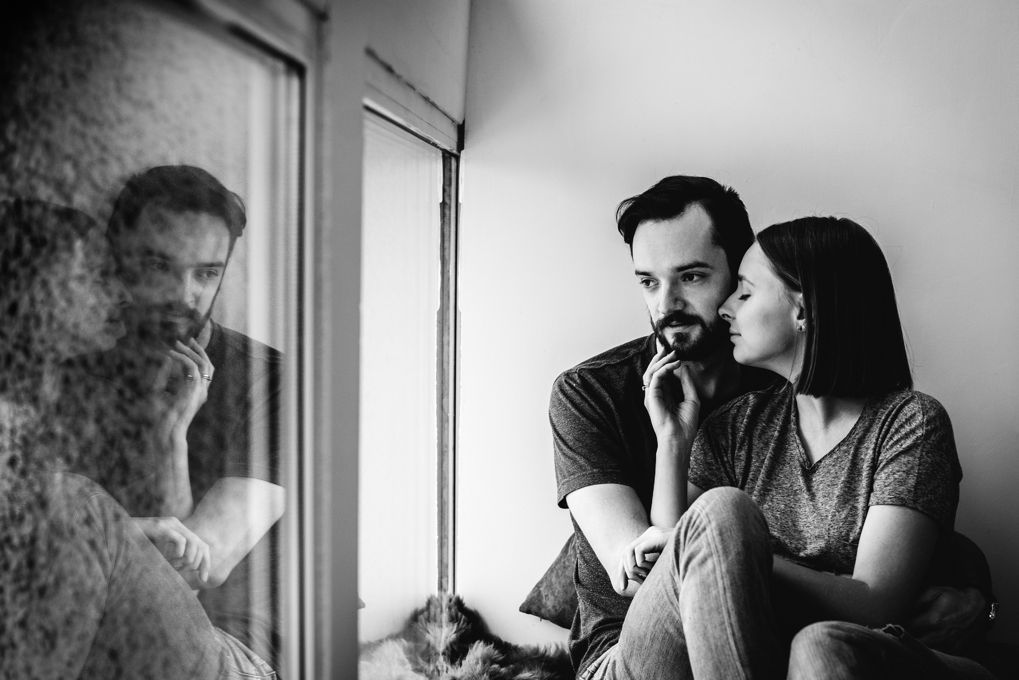 A monochrome image of a couple sitting together by a window. The woman is leaning on the man, who is gazing out of the window thoughtfully. Their reflection is seen on the glass, adding depth to the intimate and contemplative scene.