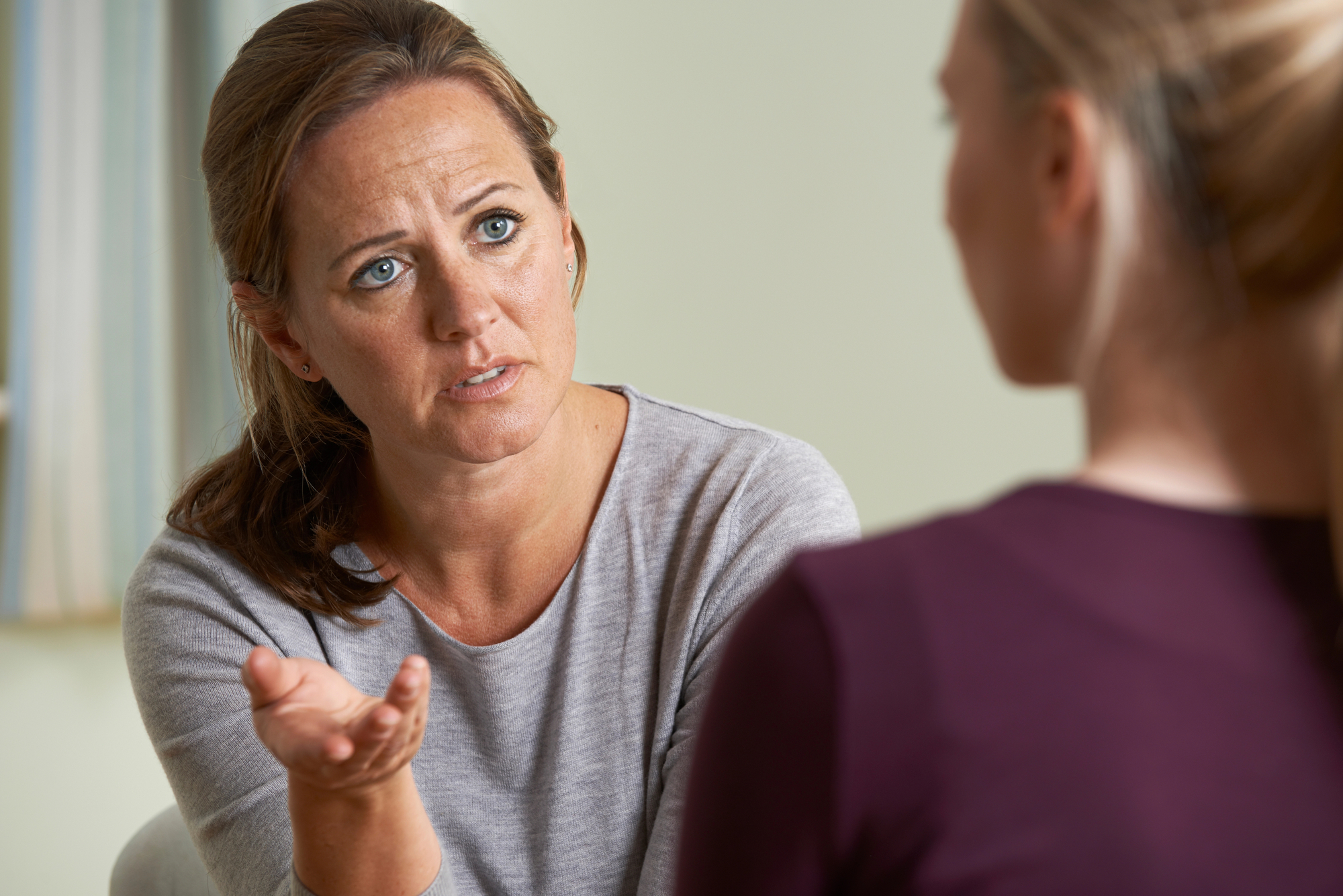 Two women are sitting and conversing. The woman on the left has a concerned expression and is gesturing with her hand while speaking. She has brown hair tied back and is wearing a grey top. The woman on the right is facing away, with blonde hair and a maroon top.