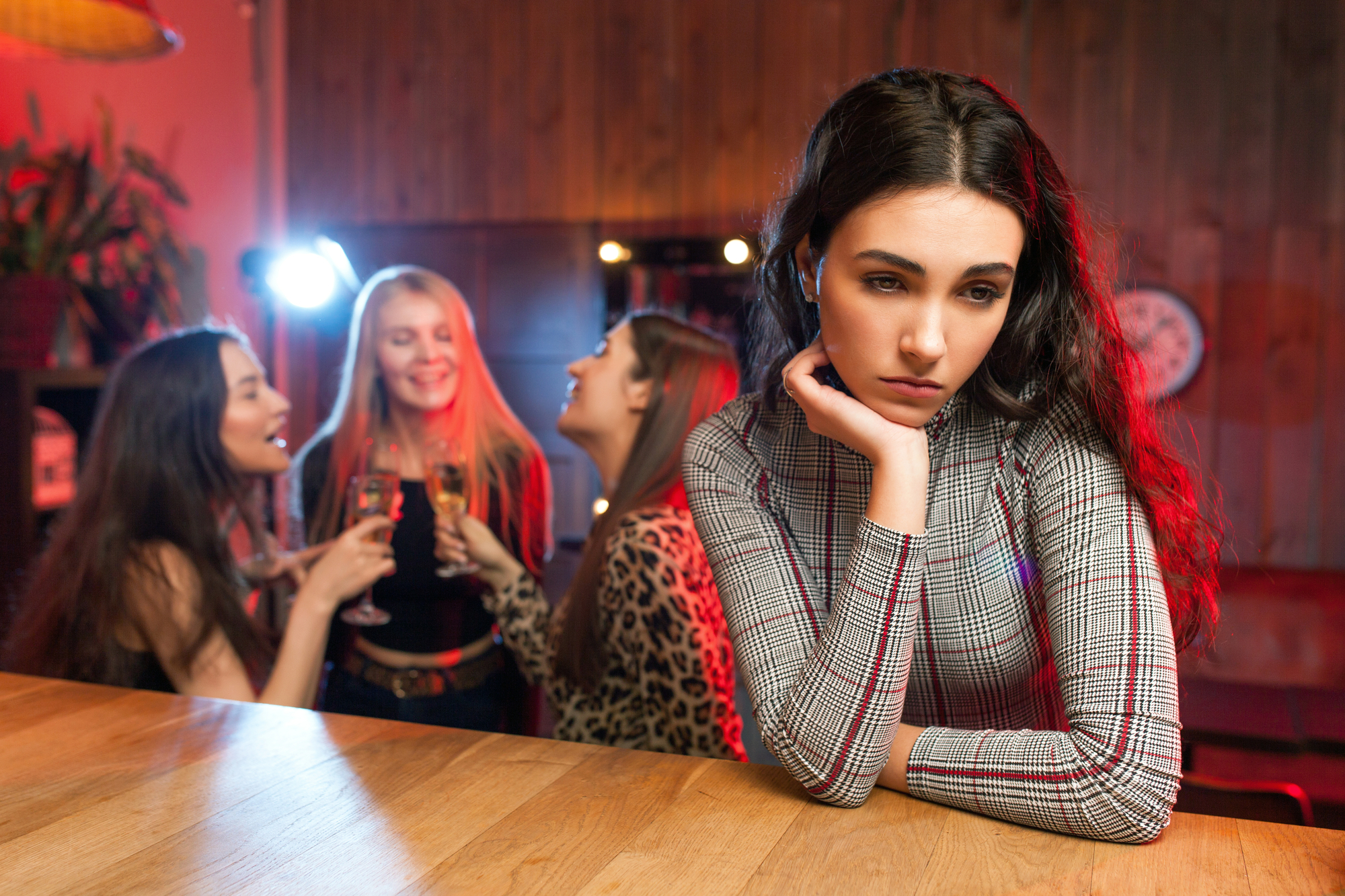 A woman with long dark hair and a plaid sweater sits alone at a bar, resting her face on her hand and looking pensive. In the background, a group of three women are enjoying themselves, smiling, and holding drinks. The setting appears to be a lively bar.