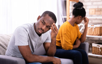 A man and woman sit on a couch, both appearing upset. The man, in a light gray shirt, leans forward with a hand on his forehead, while the woman, in a mustard yellow shirt, sits with her back to him, resting her head in her hand. The room has a brick wall and shelves.