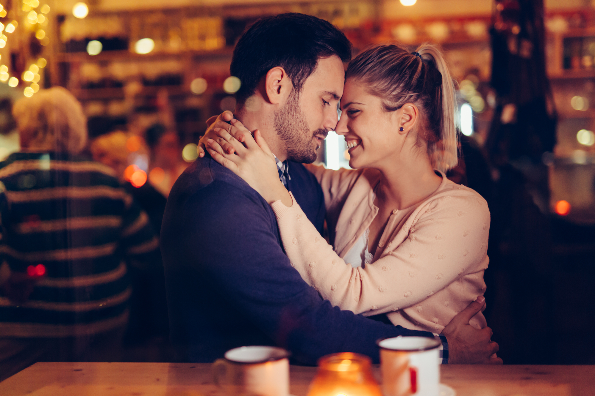 A couple sits closely together in a cozy cafe, embracing and smiling with their foreheads touching. The background is softly lit with warm lights and blurred people. Two cups of hot drinks and a candle are on the table in front of them.