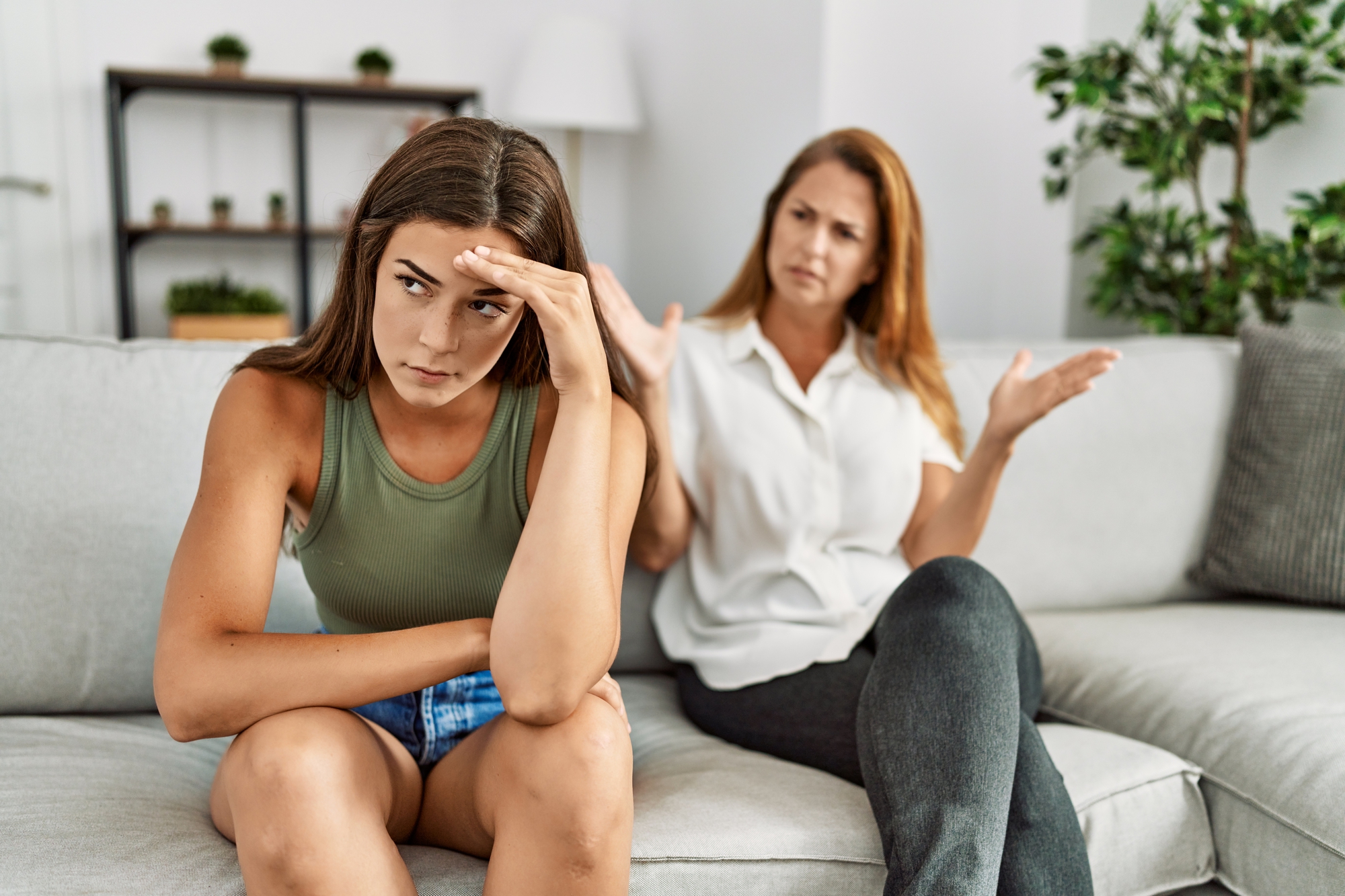 A young woman sits on a couch with a hand on her forehead, looking frustrated. An older woman beside her gestures with open hands, appearing upset or concerned. They are in a living room with plants and shelves in the background.