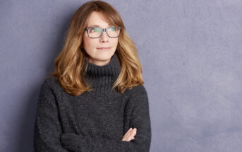 A woman with long blonde hair and wearing glasses is standing against a gray textured wall. She is dressed in a dark gray sweater and has her arms crossed, looking slightly to the side with a thoughtful expression.